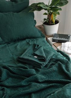 a bed with green sheets and pillows next to a potted plant on a table