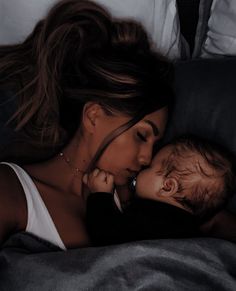 a woman laying on top of a bed next to a baby in her lap and kissing her cheek