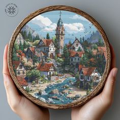 a hand holding up a cross stitch picture with houses and water in the river on it