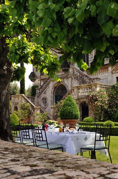 an outdoor dining area with tables and chairs in front of a large stone building surrounded by greenery