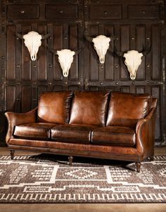 a brown leather couch sitting in front of a wooden wall with three steer heads on it