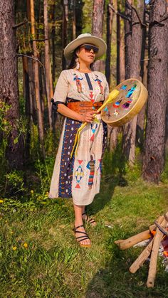 a woman wearing a hat and dress holding a fan in the grass near some trees