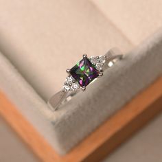 an engagement ring in a box with some diamonds on it's sides and a green mystic stone surrounded by smaller white stones