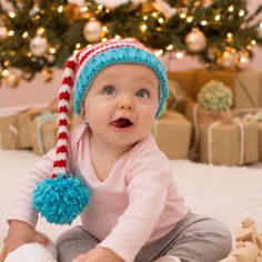 a baby sitting on the floor wearing a blue and red knitted hat with pom poms