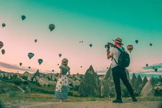 a man and woman standing on a dirt road looking at hot air balloons in the sky