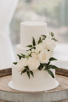 a wedding cake with white flowers and greenery on the top is sitting on a wooden barrel