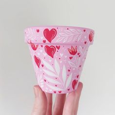 a hand holding a pink cup with hearts and leaves on the outside, in front of a white background