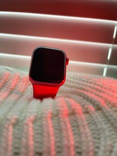 a red apple watch sitting on top of a white blanket in front of a window