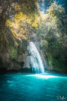 a waterfall in the middle of a river with blue water and trees around it, surrounded by lush green foliage