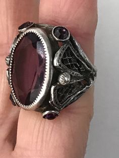 "This lovely filigreed and ornate ring has an oval shaped faceted amethyst purple color glass stone at its center. It is flanked on either side of to and bottom with small round bezel set purple glass stones. The sides have lattice shaped cut outs and the band is smooth. The face of this ring is about 3/4\" long and about 1/2\" across. It is a size 3 1/4 and is marked Sterling. Please note there are some overall signs of wear, especially to the stones, four of five of which, including the center Oval Purple Filigree Amethyst Ring, Purple Oval Filigree Rings, Oval Purple Amethyst Ring With Filigree, Purple Oval Amethyst Ring With Filigree, Oval Jeweled Ring For Gift, Oval Jeweled Rings For Gifts, Purple Oval Stone Jewelry, Purple Oval Stones Jewelry, Oval Jeweled Rings Perfect For Gifts