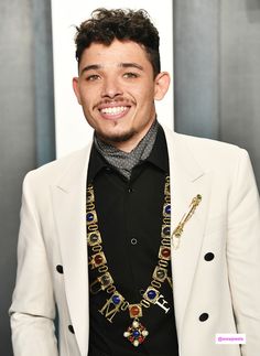 a man in a white jacket and black shirt smiles at the camera while wearing an elaborate necklace