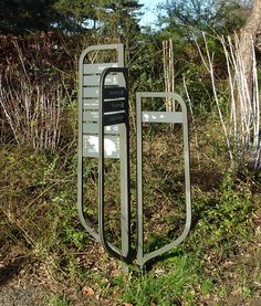 there is a metal sign in the middle of some grass and bushes with trees behind it