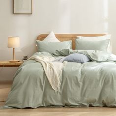 an unmade bed with grey linens in a white walled room next to a wooden headboard