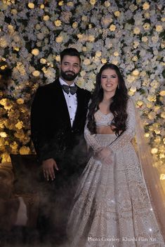 two people standing next to each other in front of a flower wall with white flowers