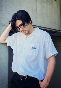 a young man wearing glasses leaning against a wall with his hand on his head and looking at the camera