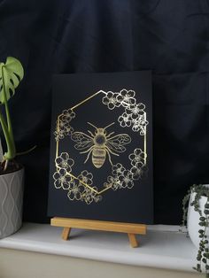 a black and gold card with a bee in the center on a wooden stand next to a potted plant