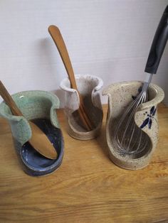 three ceramic utensils are lined up on a wooden counter top, one has a whisk in it and the other is empty