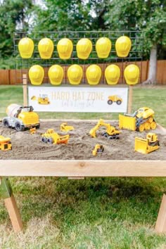 a wooden table topped with yellow toys and construction vehicles on top of sand in front of a sign that says hard hat zone