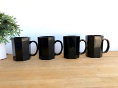 five black coffee mugs lined up on a wooden table next to a potted plant