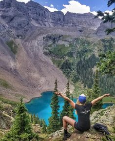 a woman sitting on top of a mountain with her arms outstretched in front of a lake