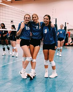 three girls in blue uniforms standing on a volleyball court with their arms around each other