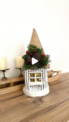 a small house with a wreath on top of it sitting on a table next to two candles