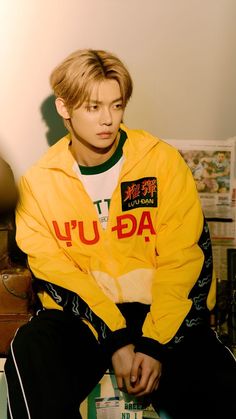 a young man sitting on top of a wooden table wearing a yellow jacket and black pants