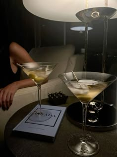 two martini glasses sitting on top of a table next to a book and a lamp