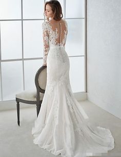 a woman in a white wedding dress standing next to a chair and looking out the window