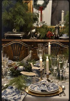 a dining room table set for christmas with blue and white dishes, silver candlesticks and greenery