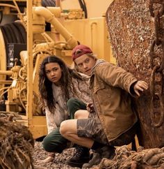 two young women crouching next to a large piece of wood in front of machinery