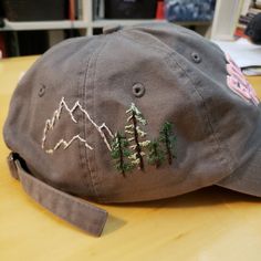 a brown hat with trees and mountains embroidered on it sitting on a table in front of a bookshelf