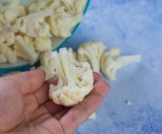 a hand holding up a piece of cauliflower next to a bowl full of chopped cauliflower