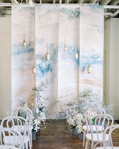 an indoor wedding venue with white chairs and floral arrangements on the wall, along with hanging lights