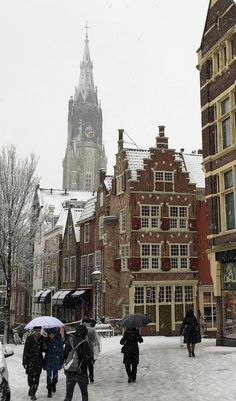 people walking in the snow with umbrellas near buildings and a clock tower behind them