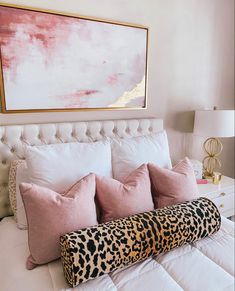 a white bed with pink pillows and a leopard print pillow on the headboard in front of a painting