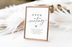 an open seating sign sitting on top of a table next to white flowers and feathers