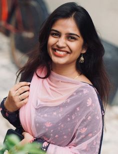 a smiling woman wearing a pink sari and carrying a black purse in her hand