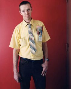 a man in a yellow shirt and tie standing against a red wall with his hands in his pockets