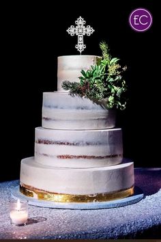 a three tiered cake with greenery on top sits on a table next to a lit candle