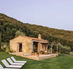 a stone house with lawn chairs in the foreground and an olive tree covered hillside in the background