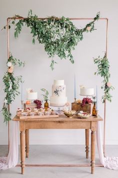 a table topped with cakes and candles covered in greenery next to a wall hanging from the ceiling