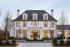 a large white house with lots of windows and bushes in the front yard at dusk