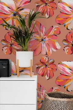 a white cabinet sitting next to a pink wall with flowers on it and a potted plant