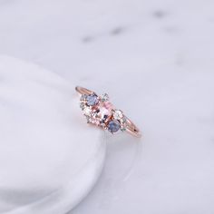 three stone ring sitting on top of a white marble plate with a pink and blue diamond in the middle