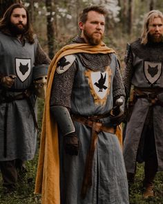 three men dressed in medieval clothing standing next to each other on grass covered ground with trees behind them