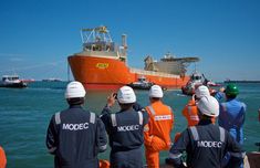 several men in orange and blue work suits watch as a large boat passes by