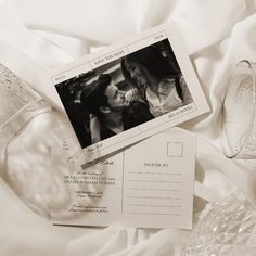 an envelope and postcard are laying on a bed with white sheets, glass vases and wine glasses