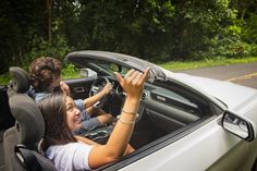 two people sitting in the driver's seat of a car, one holding up her hand