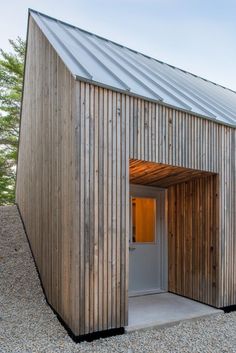 a wooden building with a metal roof and door on gravel ground in front of trees